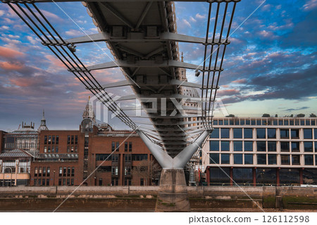 View of the Millennium Bridge's suspension cables and modern design, with St. Paul's Cathedral dome in the background under a colorful sky. 126112598