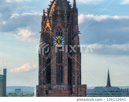 The Gothic style tower of Frankfurt Cathedral with intricate details, a gold accented clock, and modern buildings in the background under a cloudy sky. 126112641