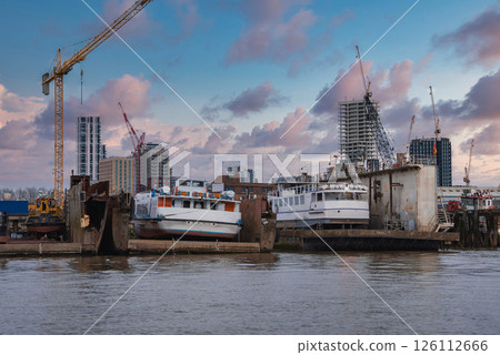 A London riverside shipyard with two white boats on platforms, surrounded by cranes and modern high rise buildings under construction at sunset. 126112666