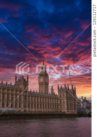 Elizabeth Tower, part of the Palace of Westminster, illuminated by a vibrant sunset. The River Thames reflects the colorful sky, with a crane visible nearby. 126112717