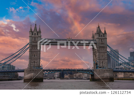 Tower Bridge spans the River Thames in London, featuring intricate towers, suspension cables, and a colorful sky. Modern buildings surround the historic landmark. 126112734