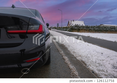 Rear view of a black BMW G20 320 with visible taillights and exhaust pipe, parked on a snowy road near houses with snow covered roofs and hedges. 126112758