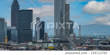 Frankfurt skyline featuring Grand Tower and Messeturm, with traditional and modern buildings, a red crane, and rolling hills under a cloudy sky. 126112765
