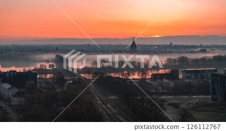 Serene sunrise over Riga, Latvia, featuring St. Peter's Church spire, mist covered Daugava River, a railway bridge, and urban structures in soft light. Serene sunrise over Riga, Latvia, featuring St. Peter's Church spire, mist covered Daugava River, a railway bridge, and urban structures in soft light. 126112767