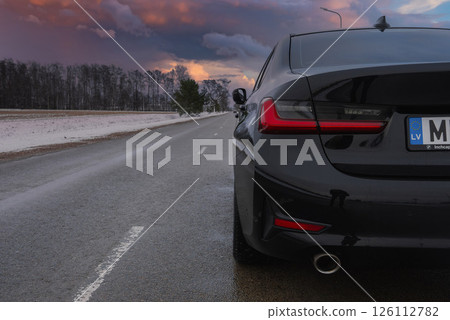 Rear view of a black BMW G20 320 with sharp taillights and exhaust pipe, parked on a wet road. Snowy roadside and bare trees under an overcast sky. 126112782