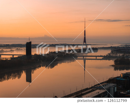 Serene sunrise over Riga, Latvia, featuring the Riga TV Tower and its reflection in the Daugava River, with a bridge and modern high rise visible. Serene sunrise over Riga, Latvia, featuring the Riga TV Tower and its reflection in the Daugava River, with a bridge and modern high rise visible. 126112799