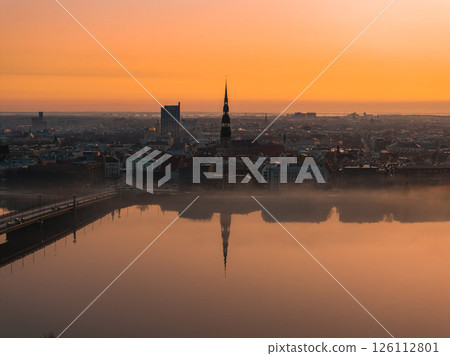Serene sunrise over Riga, Latvia, featuring St. Peter's Church spire, calm Daugava River reflections, a bridge, and a mix of historic and modern buildings. Serene sunrise over Riga, Latvia, featuring St. Peter's Church spire, calm Daugava River reflections, a bridge, and a mix of historic and modern buildings. 126112801
