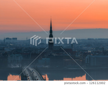 Serene sunrise over Riga, Latvia, featuring St. Peter's Church spire, a bridge over the Daugava River, and reflections in calm water with morning mist. Serene sunrise over Riga, Latvia, featuring St. Peter's Church spire, a bridge over the Daugava River, and reflections in calm water with morning mist. 126112802