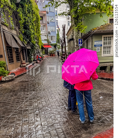 Couple holding pink umbrella strolls along a wet cobblestone street with greenery. High quality photo 126113554