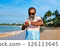 Senior ethnic man with beard uses smart watch on sunny beach. Modern tech tracks health stats. Wireless earbuds connected. Older generation enjoying gadgets. Tech-savvy grandpa into wearable devices. 126113645