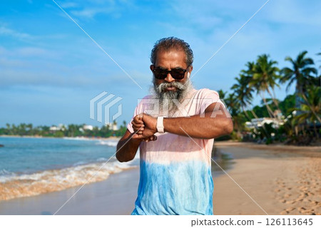 Senior ethnic man with beard uses smart watch on sunny beach. Modern tech tracks health stats. Wireless earbuds connected. Older generation enjoying gadgets. Tech-savvy grandpa into wearable devices. 126113645