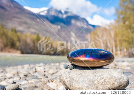 Steel tongue drum resting on a stone by the river in the Altai mountains. Concept of elemental harmony, raw sound energy, and nature as a sacred space for music and silence 126118003