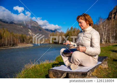 Woman playing a steel tongue drum in nature with a mountain river and Altai landscape in the background. Concept of harmony, mindfulness, and connection with nature 126118004