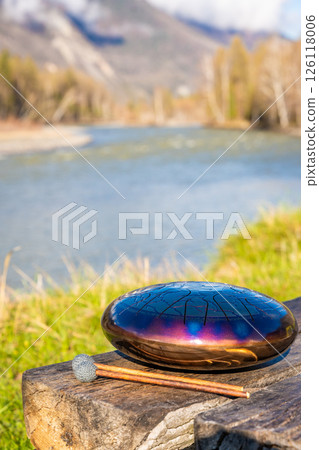 Steel tongue drum and mallets resting on a wooden bench with mountains, river, and forest in the background. Concept of sacred stillness, sound meditation, and harmony with nature 126118006