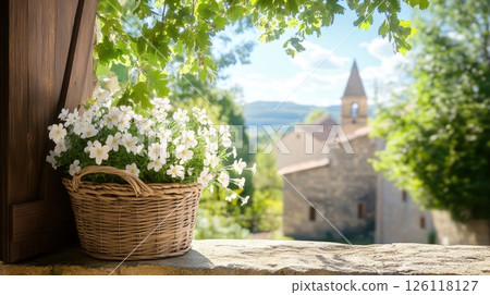 Basket of white flowers on stone windowsill with sunlit leaves and medieval French village in background featuring church spires and wooden doors, rustic summer concept 126118127