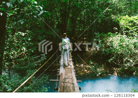 Hiker crosses a rope bridge over a turquoise river in a lush forest. 126118304