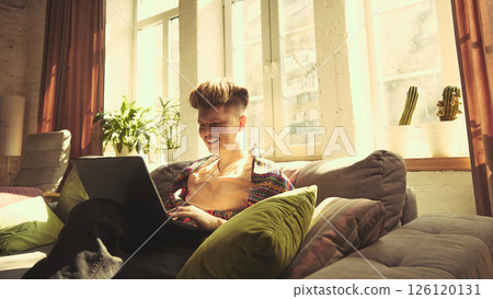 Man in colorful shirt smiling and working on laptop, sitting on grey sofa near window in living room. Man in colorful shirt smiling and working on laptop, sitting on grey sofa near window in living room. 126120131