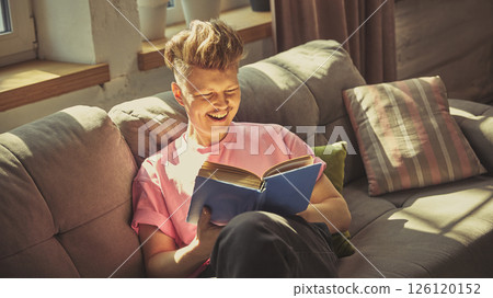 Man in pink shirt smiles joyfully while reading blue book on cozy sofa under daylight in living room 126120152