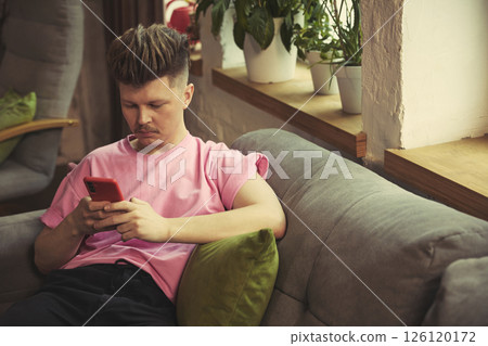 Focused man with smartphone sitting by bright window on comfortable sofa at home 126120172