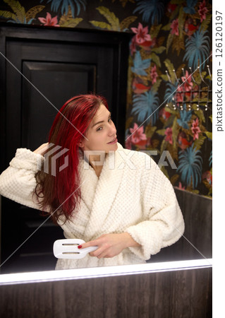 Woman with red hair, in bathrobe brushing hair looking in mirror in bathroom at home. Morning routine, grooming. Woman with red hair, in bathrobe brushing hair looking in mirror in bathroom at home. Morning routine, grooming. 126120197