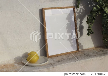 Minimalist summer still life scene. Blank wooden picture frame mockup leaning on a white textured wall. Ceramic plate, two fresh lemons, bathed in natural sunlight. Leafy shadows. Eucalyptus branches 126120867