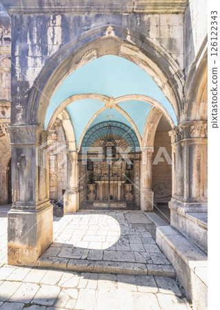 Korcula town in island of Korcula in Adriatic sea in Croatia, Europe. Stone arches frame a gated chapel with a blue ceiling, casting light and shadow on the stone-paved ground. 126122343