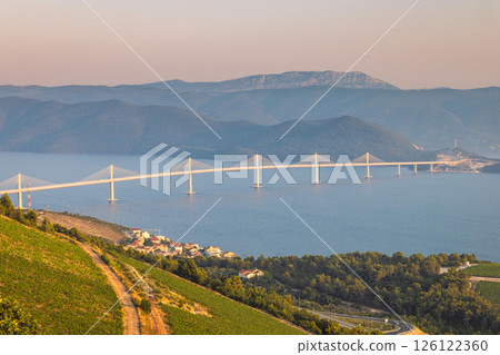 The Peljesac Bridge to Peljesac peninsula in Croatia, Europe. Cable-stayed bridge gracefully spanning a body of water, with mountains in the background, as seen from a vineyard hillside. The Peljesac Bridge to Peljesac peninsula in Croatia, Europe. Cable-stayed bridge gracefully spanning a body of water, with mountains in the background, as seen from a vineyard hillside. 126122360