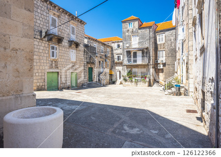 Korcula town in island of Korcula in Adriatic sea in Croatia, Europe. Old town square with stone buildings, cobblestone ground, green doors, stairs, and a vintage well under a bright blue sky. 126122366