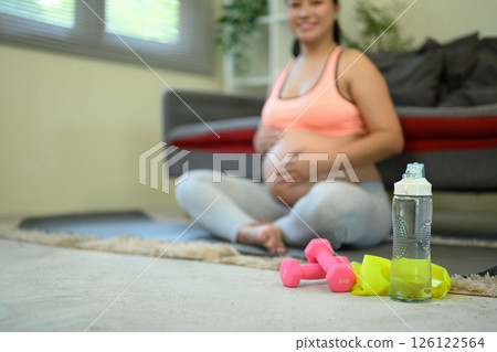 Pregnant woman sitting on the floor with a bottle of water and two dumbbells in front of her 126122564