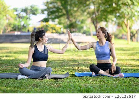 Female friends sitting on yoga mats, giving each other high five after workout in the park 126122617