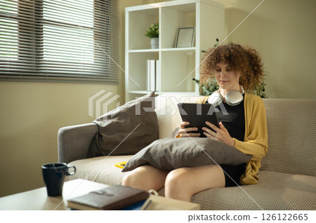 Young woman relaxing at home with headphones around her neck browsing on digital tablet Young woman relaxing at home with headphones around her neck browsing on digital tablet 126122665