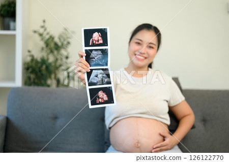 Happy expectant mother proudly showing ultrasound photos while gently touching her bare belly 126122770