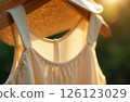 Women's beige sundress and straw hat hanging on wooden hanger on green background, glare of sun 126123029