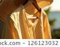 Woman Shirt Dress And Straw Hat Hanging On Hanger In Sun In Background In Blur Sea And Greenery 126123032