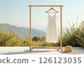 A white dress hangs on a wooden clothes hanger, a straw hat lies on the floor, against the background of nature 126123035