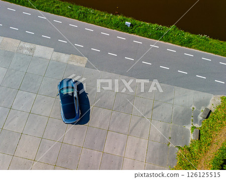 Aerial view of a single parked car on a large empty concrete lot next to a road and canal, with green grass and clear road markings under bright daylight. Aerial view of a single parked car on a large empty concrete lot next to a road and canal, with green grass and clear road markings under bright daylight. 126125515