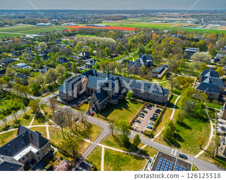 Aerial view of a peaceful suburban neighborhood with tree-lined streets, modern and traditional buildings, and surrounding farmland stretching into the horizon under a clear sky. 126125518