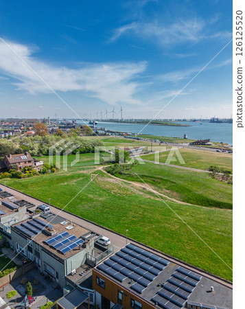 Aerial view of a coastal town with green parks, residential areas, a riverfront road, and busy shipping lanes. Wind turbines and industrial port facilities are visible across the water. Aerial view of a coastal town with green parks, residential areas, a riverfront road, and busy shipping lanes. Wind turbines and industrial port facilities are visible across the water. 126125520