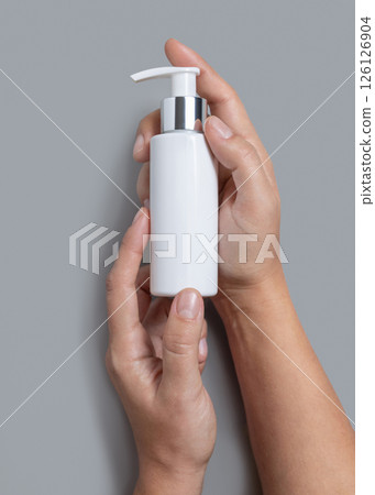 Female hands holding white cosmetic pump bottle against grey close up, mockup, copy space 126126904