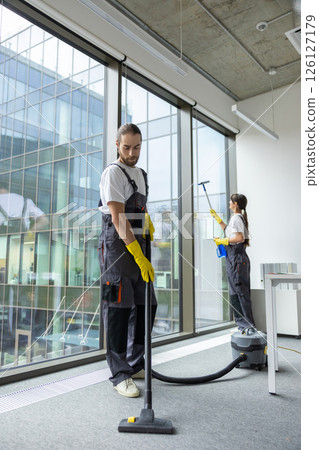 Young man in uniform vacuum cleaning the floor in the conference room 126127179