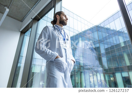 Male young doctor in white lab coat standing near the window 126127237