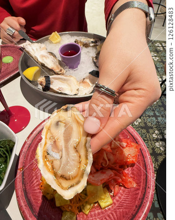 Woman's hand holding oyster above vibrant plate with pickled vegetables during gourmet meal with seafood and wine. Concept of dining, seafood indulgence, flavor fusion Woman's hand holding oyster above vibrant plate with pickled vegetables during gourmet meal with seafood and wine. Concept of dining, seafood indulgence, flavor fusion 126128483