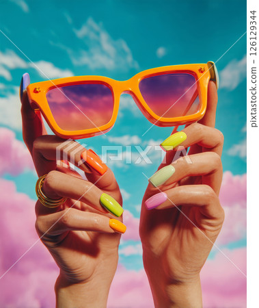 Y2K hands with colorful nails holding orange sunglasses against pink blue sky Y2K hands with colorful nails holding orange sunglasses against pink blue sky 126129344