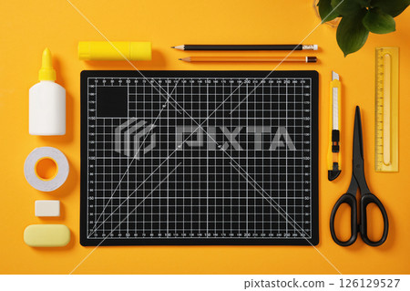 A black cutting mat with white grid lines sits on a bright yellow background surrounded by neatly laid out office supplies: pencils, glue, tape, scissors, cutter, erasers and ruler. 126129527