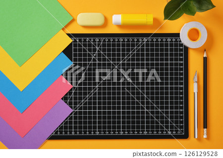 A black cutting mat with white grid lines sits on a bright yellow background surrounded by neatly laid out office paper art supplies: pencil, glue, tape, cutter, eraser and paper. 126129528