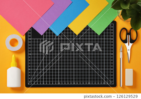 A black cutting mat with white grid lines sits on a bright yellow background surrounded by neatly laid out art hobby supplies: pencil, glue, tape, scissors, cutter, eraser and paper 126129529