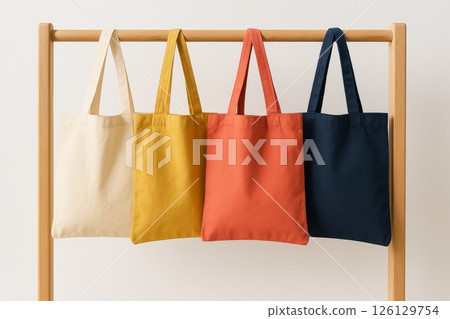 Four plain canvas tote bags in cream, yellow, orange, and navy colors hanging on wooden rack, neutral background, studio shot 126129754
