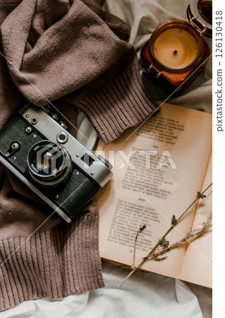 Vintage film camera with an old book wrapped in a warm knitted sweater and an extinguished candle next to it. View from above on a flat surface 126130418