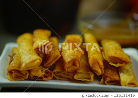 Golden-brown crispy Indonesian risoles or spring rolls stacked on a white plate, served with a small bowl of dipping spicy peanut sauce and a toothpick, perfect for an appetizing snack or appetizer. 126131419