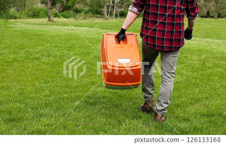 man in casual clothes holds bag with cut grass of lawn mower after mowing lawn with at own backyard. Husband takes care of garden. Modern gasoline garden equipment. Landscaping work at home 126133168
